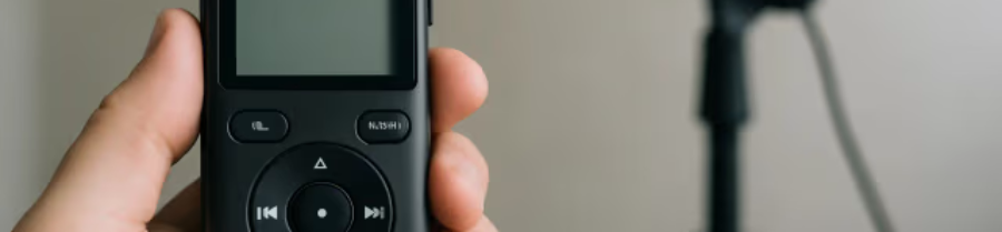 Hand holding a digital voice recorder with a studio microphone blurred in the background.