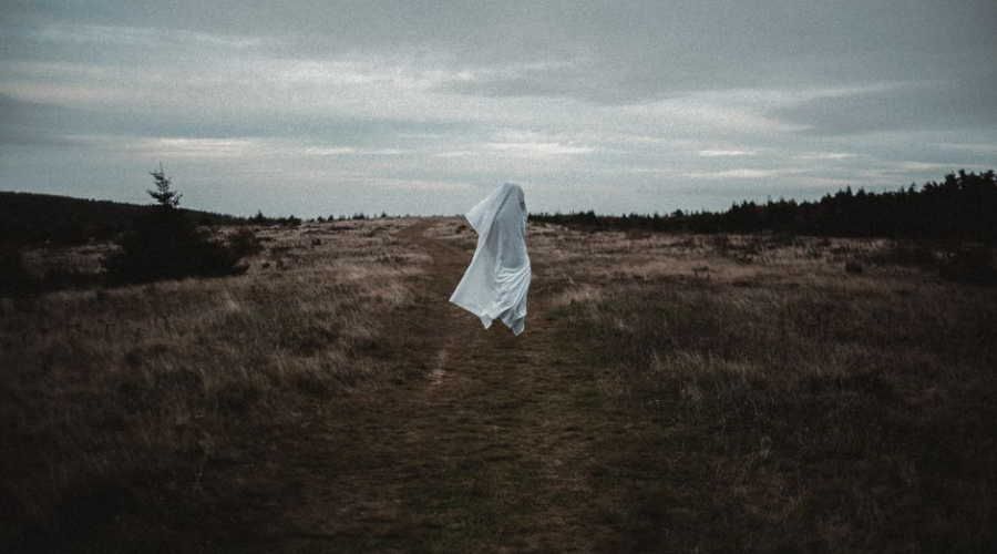 A ghostly figure in white floats over a desolate grassy field beneath a cloudy sky.