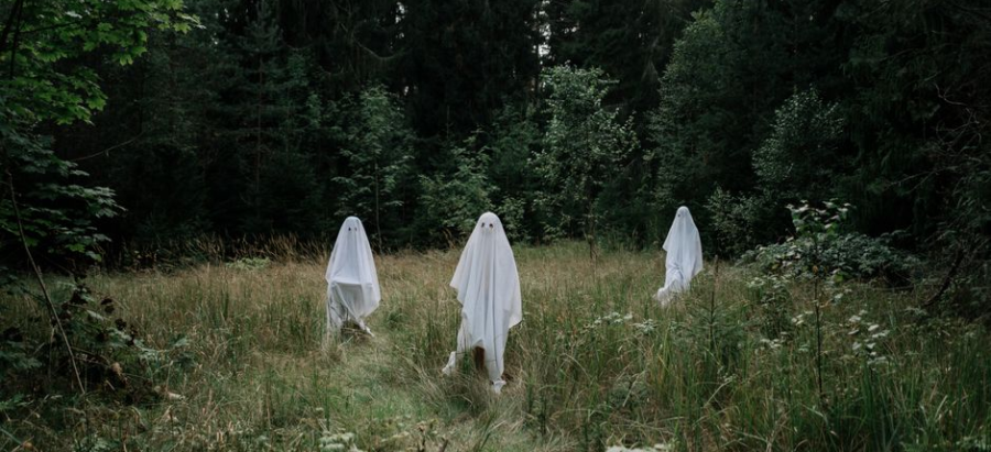 Three people dressed as ghosts in white sheets stand in a forest clearing.