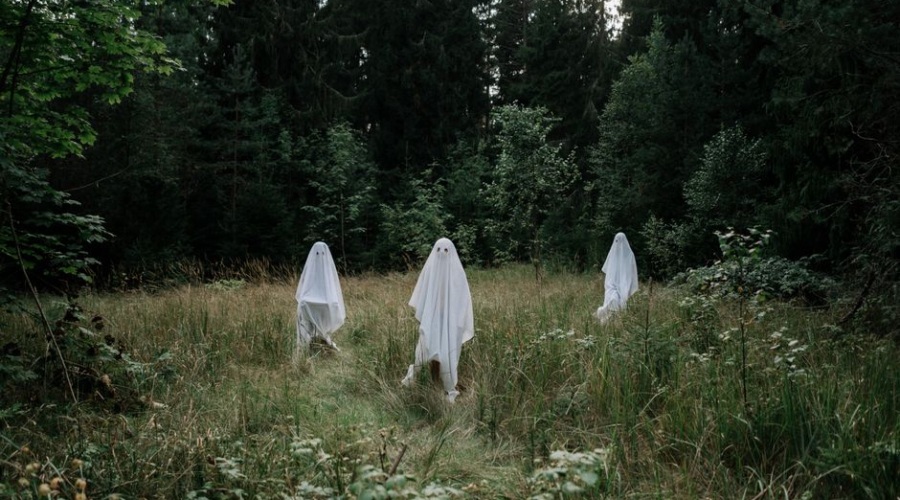 Three people dressed as ghosts in white sheets stand in a forest clearing.