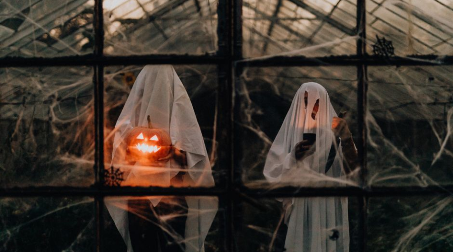 Two sheet ghosts with jack-o'-lanterns in a cobweb-filled spooky greenhouse