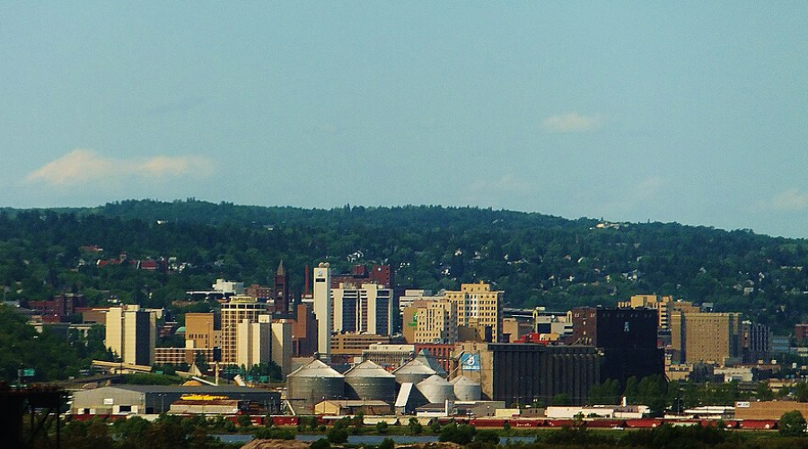 Panoramic Duluth skyline with modern buildings, domed arena, and wooded hills under a clear blue sky.