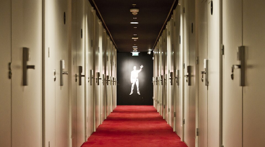 Dimly lit hotel hallway with red carpet and closed doors leads to a glowing figure in the distance