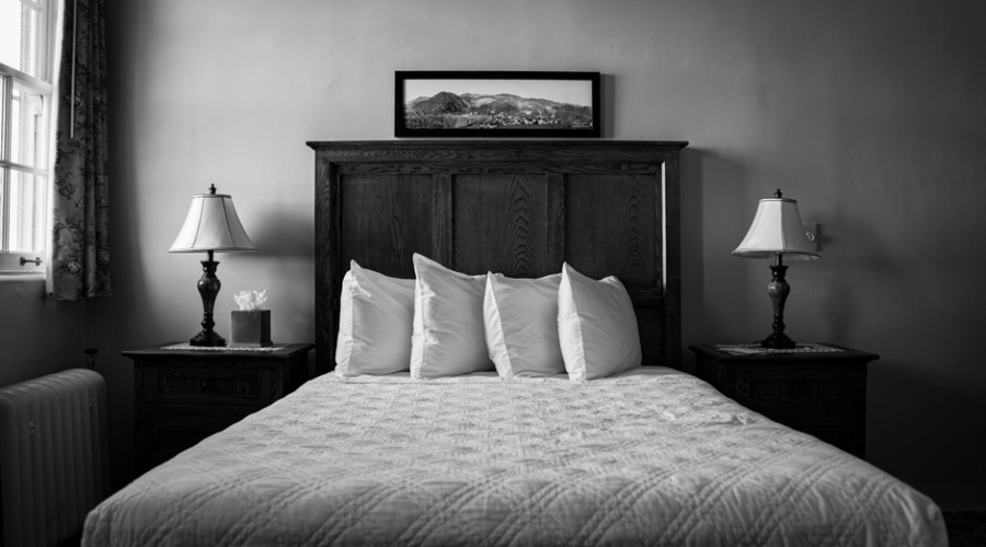 Black and white photo of a neatly made bed with four pillows and two bedside lamps in a cozy bedroom