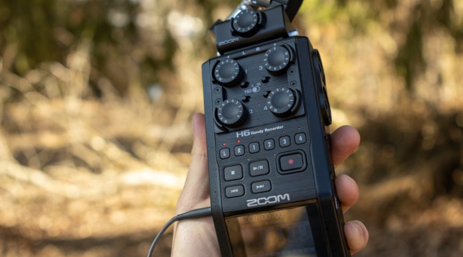 Hand holding a Zoom H6 audio recorder with microphones in a forest.